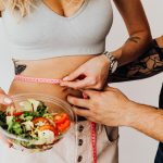 A couple measuring waistline with a tape and holding a fresh salad bowl, focusing on fitness and weight loss.
